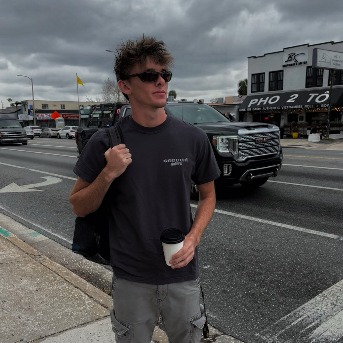 Man standing on a street corner holding a coffee cup, with a dark and cloudy sky.